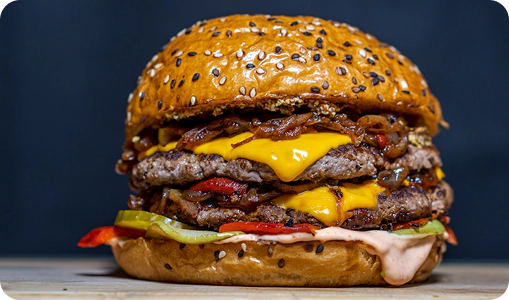 A double cheeseburger with sesame seed bun, two beef patties, melted cheese, lettuce, pickles, grilled onions, red peppers, and sauce.